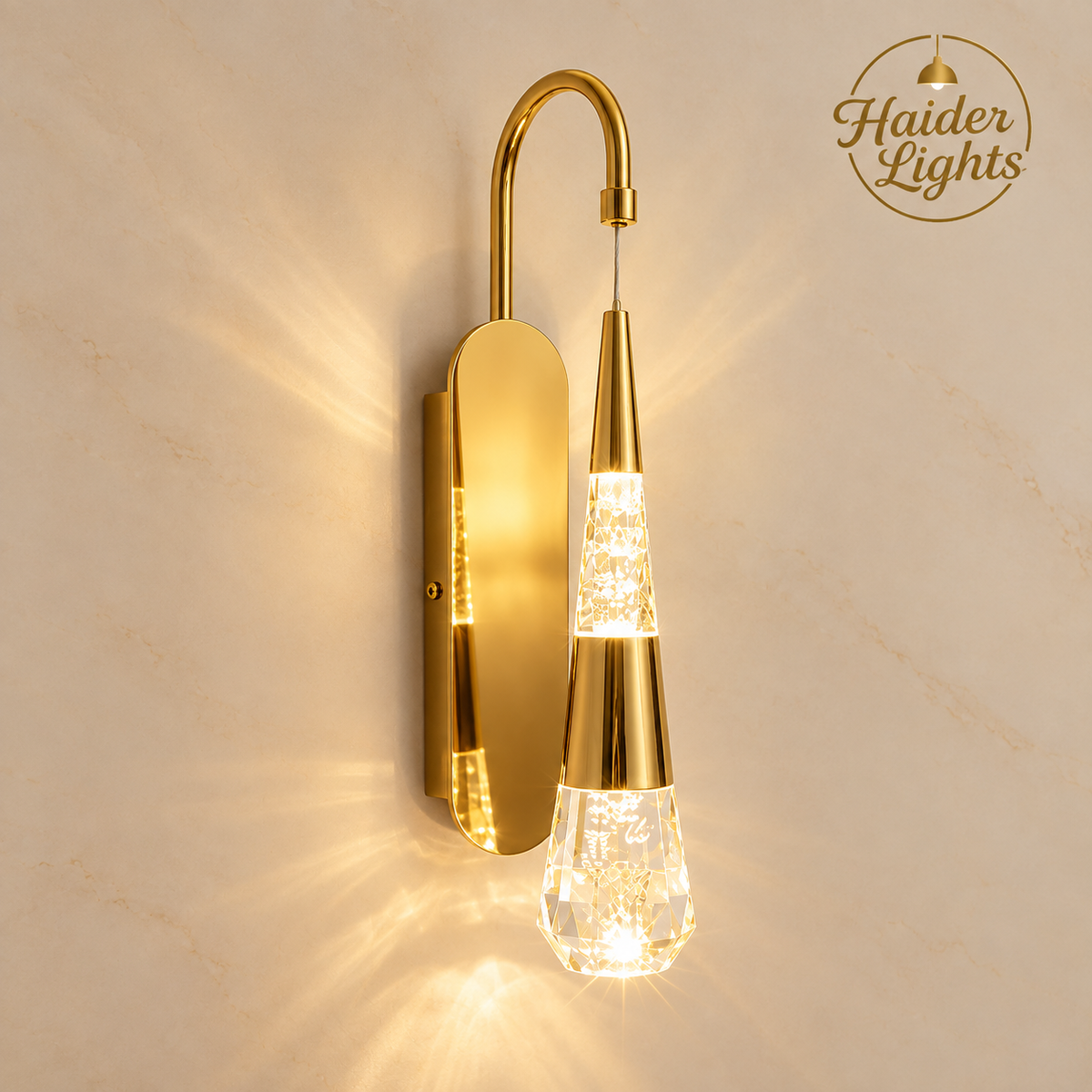 A modern wall-mounted sconce light fixture in polished gold with a clear faceted glass teardrop shade, shown illuminated against a beige marble wall. The brand logo 'Haider Lights' is visible in the top right corner.