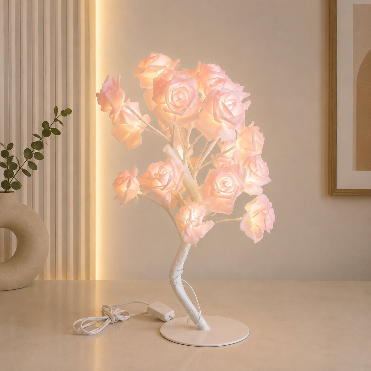 A decorative table lamp shaped like a flowering tree with glowing pale pink roses on a white curved base, shown on a tabletop in a softly lit interior setting.