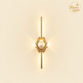 A modern gold wall sconce with a vertical rod, a central lotus-like gold frame, and a clear faceted crystal prisma, shown against a warm cream background with a brand logo in the top right.