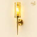 A modern wall sconce light fixture with a cylindrical amber-tinted glass shade, a visible filament bulb, and a brushed brass metal mount and spear-shaped stem, shown against a warm off-white wall. The Hauder Lights logo is visible in the top right corner.