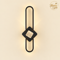 A modern wall sconce light fixture with a black metal U-shaped frame, a dark wood diamond accent, and a textured gold square in the center, emitting a warm glow against a beige background. The Haider Lights logo is visible in the top right corner.
